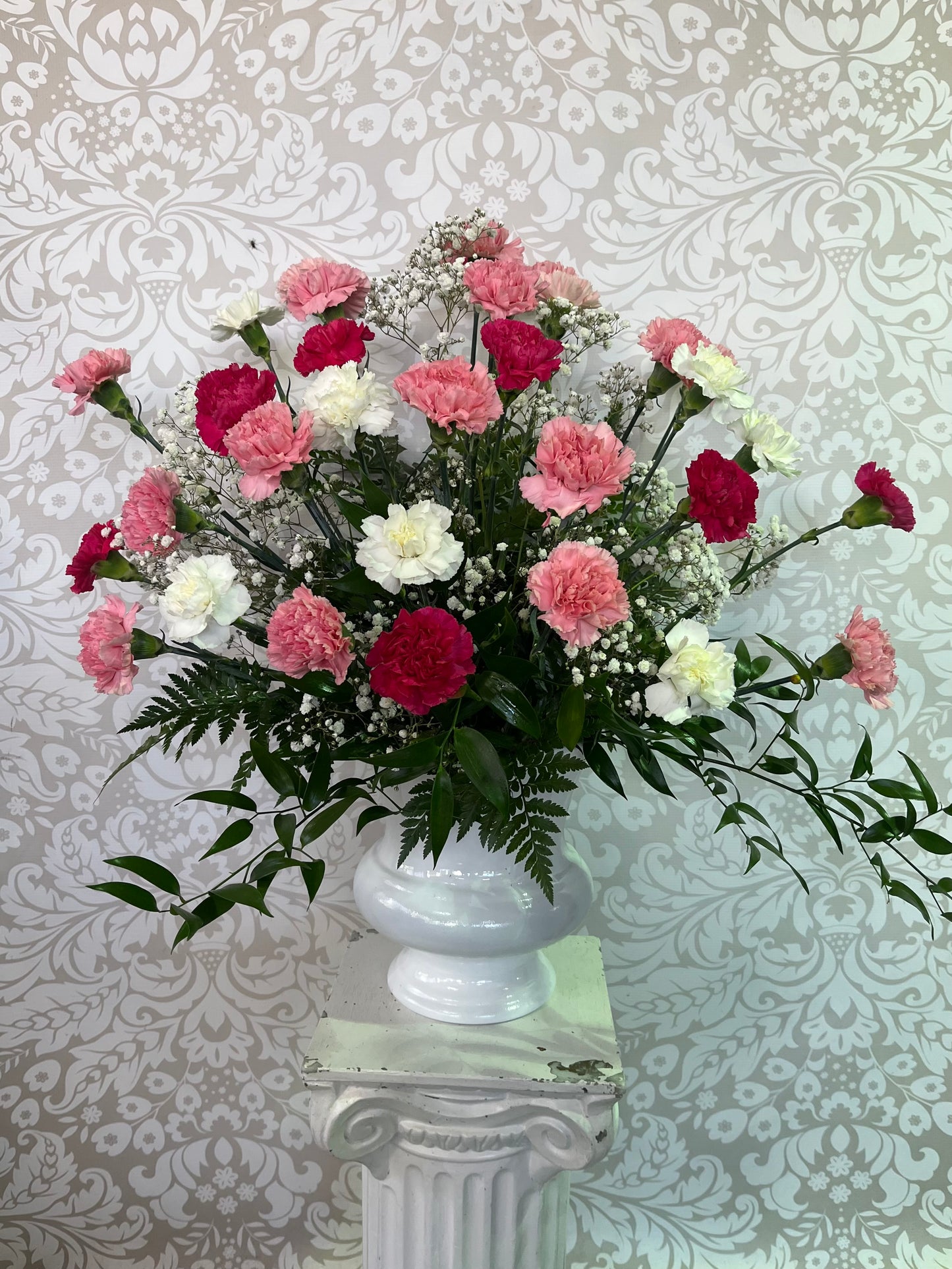 A large floral arrangement featuring pink, red, and white carnations mixed with green foliage and baby's breath, presented in a white ceramic urn on a white pedestal against a damask wallpaper background.