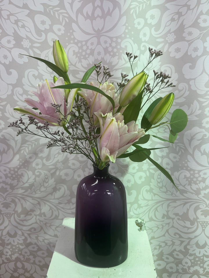 A dark purple glass vase filled with pink lilies and green buds, set on a white pedestal against a gray damask wallpaper background.
