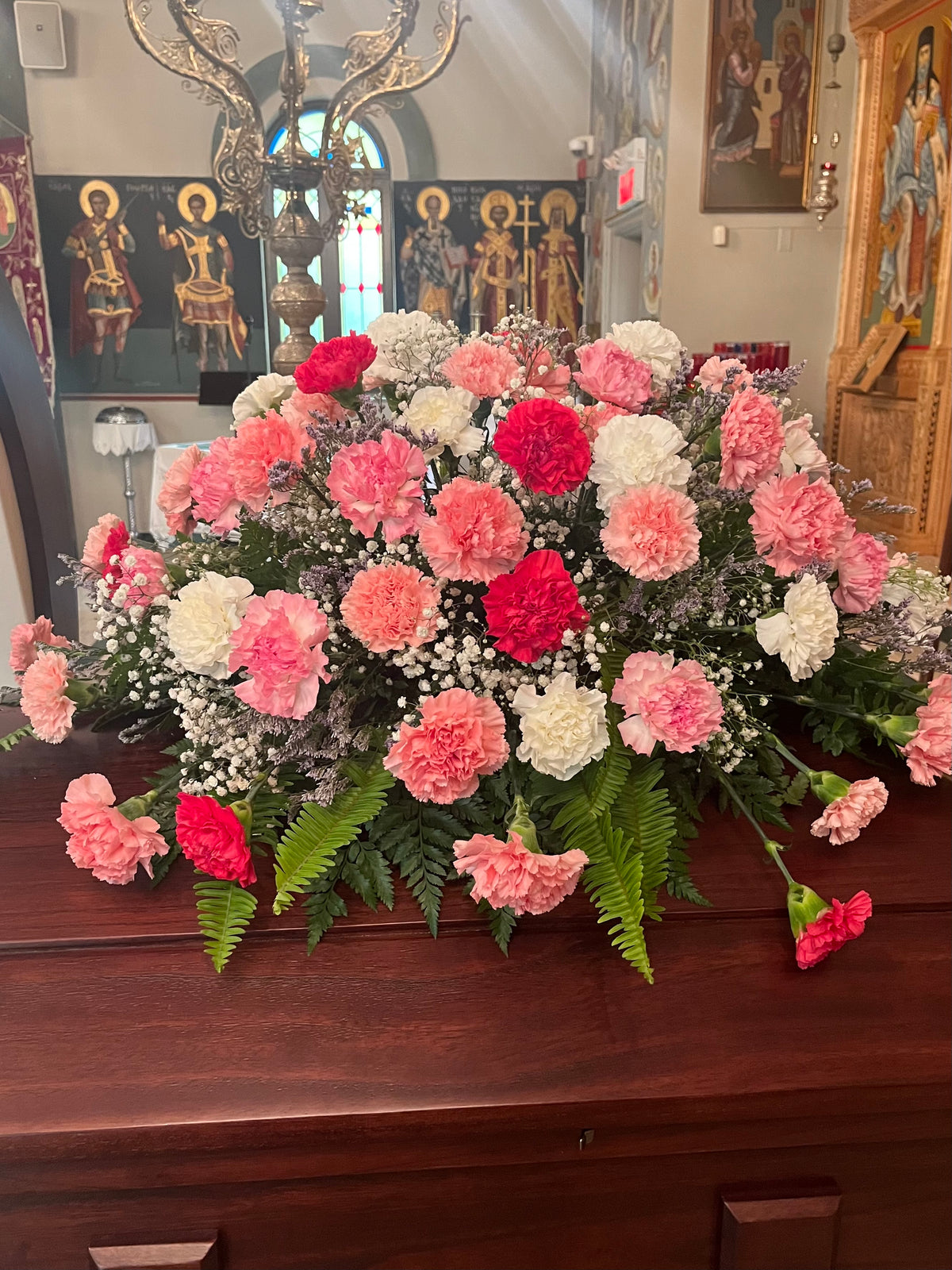 A large floral arrangement of pink, red, and white carnations with green ferns and baby's breath, displayed on a dark wooden casket in a church interior with religious icons in the background.
