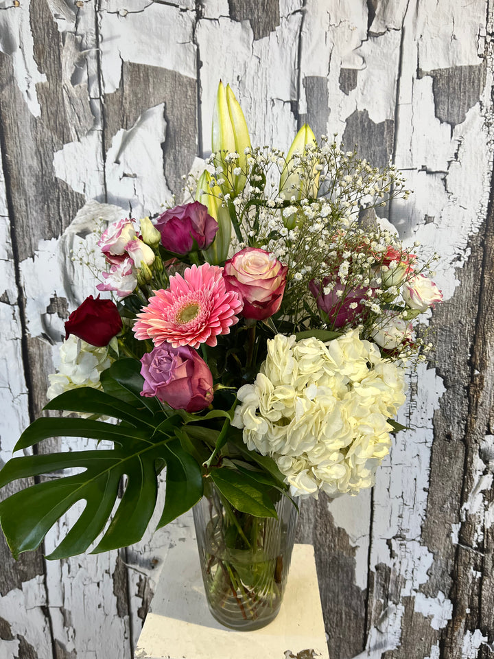 A large mixed flower arrangement in a vase, featuring a variety of flowers in shades of pink, white, and green, with a rustic wooden background.