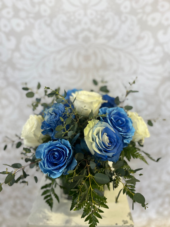 An arrangement of blue and white roses in a clear 5 inch cube vase, with a decorative pattern in the background.