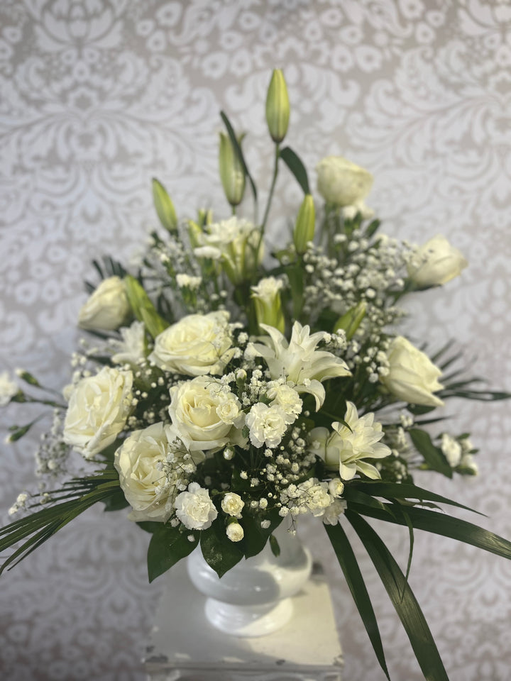 A floral arrangement featuring a classic mix of white lilies, roses, and carnations with green leaves and white flower buds.