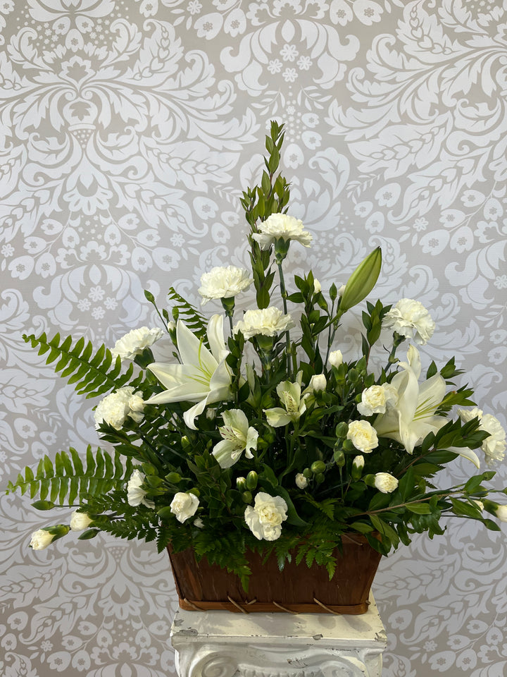 A floral basket arrangement with a variety of green and white flowers, placed on a white surface with a patterned wall in the background.