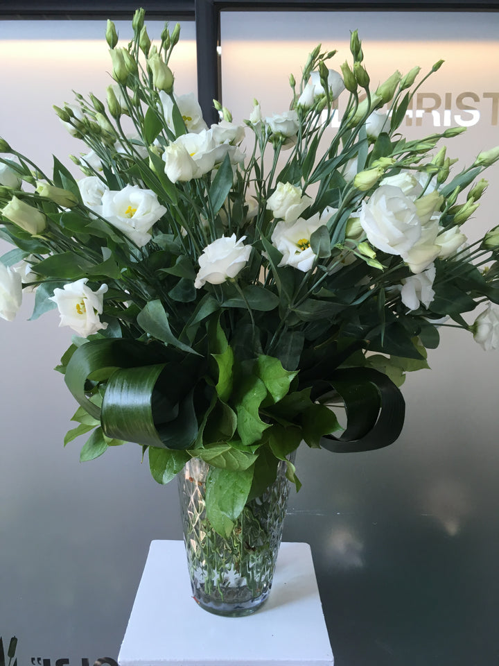 A clear glass vase containing an arrangement of white lisanthus flowers with green leaves.