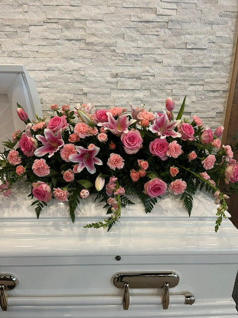 Pink casket spray with roses, lilies, carnations, and greenery on a white casket