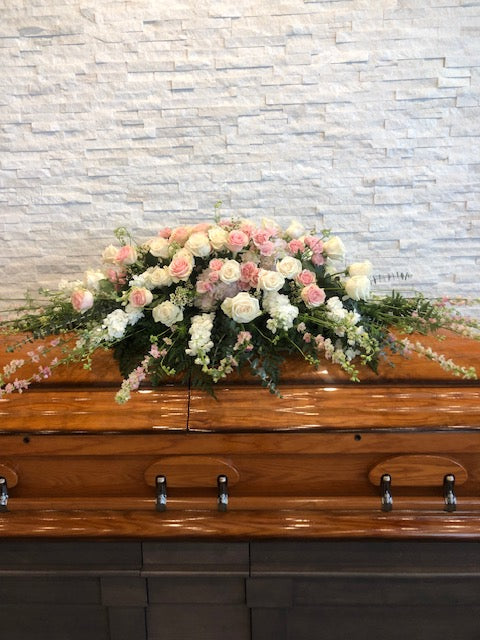 Soft pink and white casket spray with roses and greenery on a wooden casket
