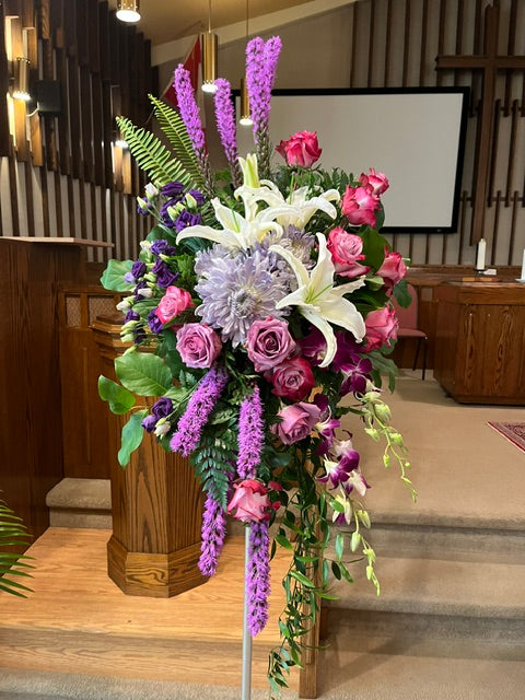 Standing sympathy floral tribute with white lilies, purple flowers, pink roses, and cascading greenery
