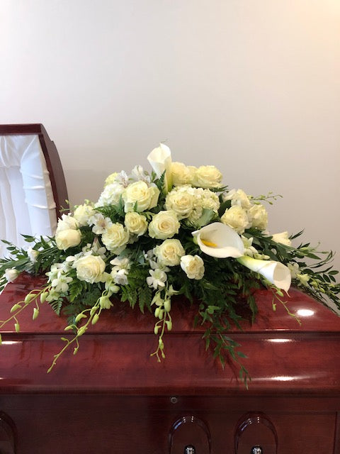 White casket spray with roses, calla lilies, and greenery on a dark wood casket